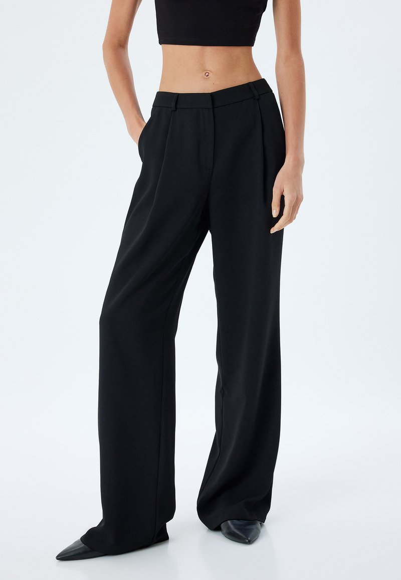 Zwarte wijde broek van zachte stof, met plooien en een gladde tailleband. Gecombineerd met puntige zwarte schoenen voor een gestroomlijnde uitstraling.