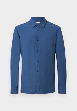 Camisa azul de manga larga con cuello, seis botones visibles y un sutil logo bordado en el pecho, mostrada sobre un fondo sencillo.