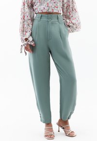 Groene, hooggetailleerde broek van gladde stof, met een dubbele knoopsluiting, zijzakken en taps toelopende pijpen.