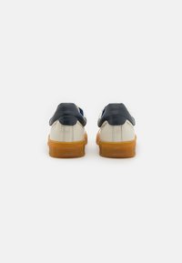 Hvite lær sneakers med en marineblå hælkappe og en gummisåle, med avrundede kanter og et merket bakparti. Teksturet overdel i lær.