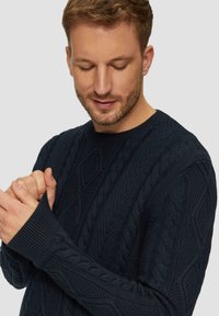 Donkerblauwe gebreide trui met een gestructureerd kabelpatroon, geribde manchetten en een ronde halslijn, gestyled met aansluitende mouwen.