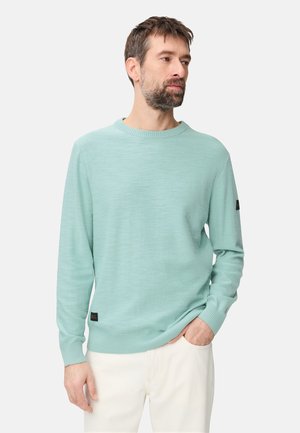 Mand med kort brunt hår og skæg iført en lyseblå sweater og hvide bukser, stående med den ene hånd i lommen og kigger til højre.