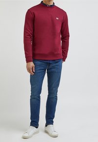 Burgundy-coloured collegepaita, jossa on pyöreä kaula-aukko, ribbaus hihansuissa ja helmassa. Valkoinen logo kauluksessa, yhdistetty sinisiin farkkuihin ja valkoisiin tennareihin.