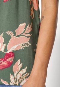 Květinová blůza s zeleným pozadím a růžovými a bordó květinami, vyrobená z měkkého materiálu, s krátkými rukávy. Detailní záběr rukávu a květinového vzoru.