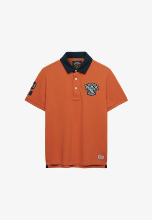 Oranje polo met korte mouwen en een marineblauwe kraag, drie witte knopen, geborduurd embleem op de linkerborst en gedetailleerde patches op de linker mouw en zoom.