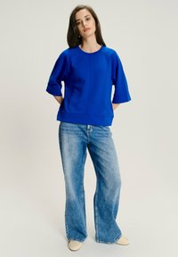 Donna in piedi con le mani dietro la schiena, indossa una maglietta blu cobalto a maniche corte, jeans larghi azzurri chiari e scarpe beige su sfondo neutro.