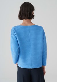 Blauwe langwerpige top met een gestructureerd gestreept patroon, ronde halslijn en een rechte zoom. De achterkant toont naaddetails. Gemaakt van zachte stof.