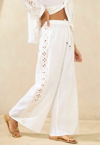 Pantalons larges blancs en tissu léger, avec des découpes florales sur les côtés, dotés d'une taille ajustable et d'une coupe décontractée.