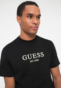 Muž s krátkymi tmavými vlasmi oblečený v čiernom tričku Guess s bielym textom "GUESS EST. 1981" vpredu, pozerajúci mierne doprava na bielom pozadí.