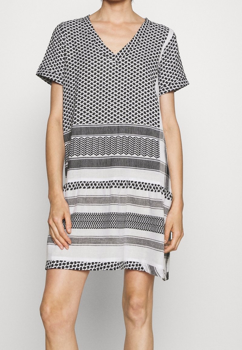 Kobieta w czarno-białej sukience z krótkim rękawem i dekoltem w kształcie litery V, zdobionej geometrycznymi i paskowanymi wzorami.