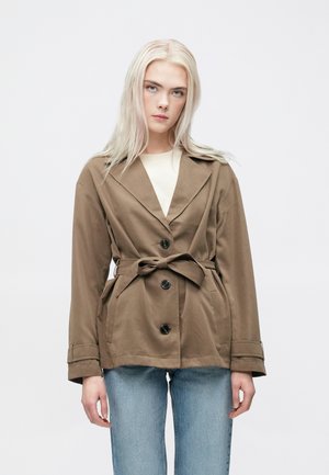 Smeđa jakna s remenom, sa postavljenim okovratnikom, prednjim gumbima i bočnim džepovima. Nosiva preko svijetle majice i uparena s svijetloplavim trapericama.