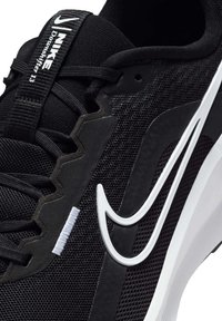 Zapatilla deportiva negra con parte superior de malla, logo de swoosh blanco y acentos texturizados. Cuenta con cordones y marca en la lengüeta.