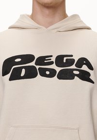 Sudadera beige con texto en negro estilizado "Pega Dor" en el pecho, llevada por una persona que se muestra desde el cuello hasta la mitad del torso.