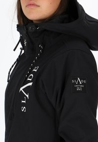 Czarna kurtka z zapięciem na zamek z przodu, regulowanym kapturem i białym logo. Posiada łatkę na rękawie z nazwą marki i szczegółami.