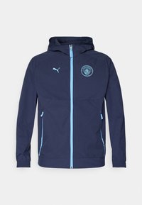 Marineblauwe hooded jack gemaakt van lichtgewicht materiaal, met een rits, zijzakken en een Manchester City-logo op de borst.