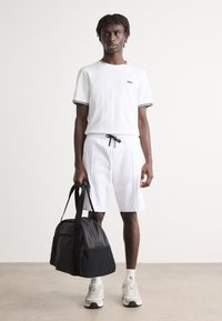 Chemise blanche à manches courtes avec bordures noires, short blanc assorti, tenant un sac de sport en tissu noir et portant des baskets grises avec des chaussettes blanches.