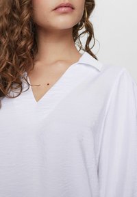 Blusa bianca con scollatura a V e maniche corte, caratterizzata da una texture stropicciata e un colletto sottile. Decorata con dettagli di cucitura minimali.