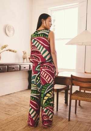 Frau steht drinnen in der Nähe eines Holztisches und Stühlen, trägt ein ärmelloses Top und weite Hosen mit großem, buntem, abstraktem Blattmuster.