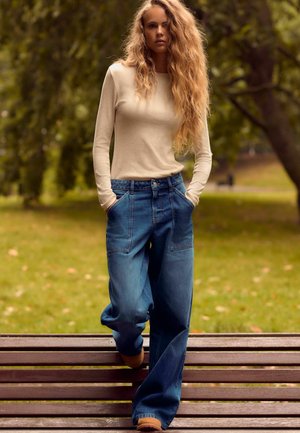 Bež pikk-sleeve topp, mis on koos vabade siniste denimite laia säärega teksadega. Tähtsad omadused on ees olevad taskud ja lõdvestatud lõige.
