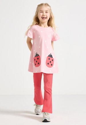 Jauna meitene rozā kleitā ar pūtīšu rakstu un lapsenīšu kabatām, sarkanas bikses un baltas sporta kurpes, smaida un soļo uz priekšu vienkrāsainā fonā.