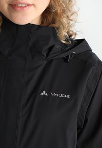 Zwarte waterdichte jas met capuchon, voorzien van een ritsluiting, gestructureerde stof en het "Vaude"-logo op de borst.