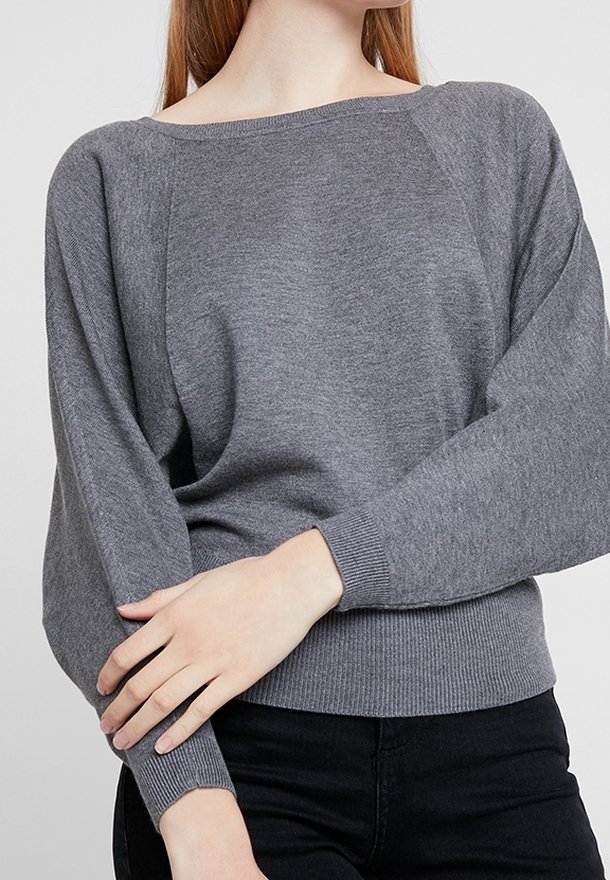 Femme portant un pull en maille gris ample avec un col bateau et un pantalon noir, les bras croisés devant à hauteur de taille.