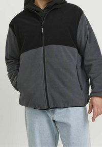Giacca in pile con sezione superiore nera e sezione inferiore grigia, chiusura a zip, due tasche laterali e vestibilità comoda.
