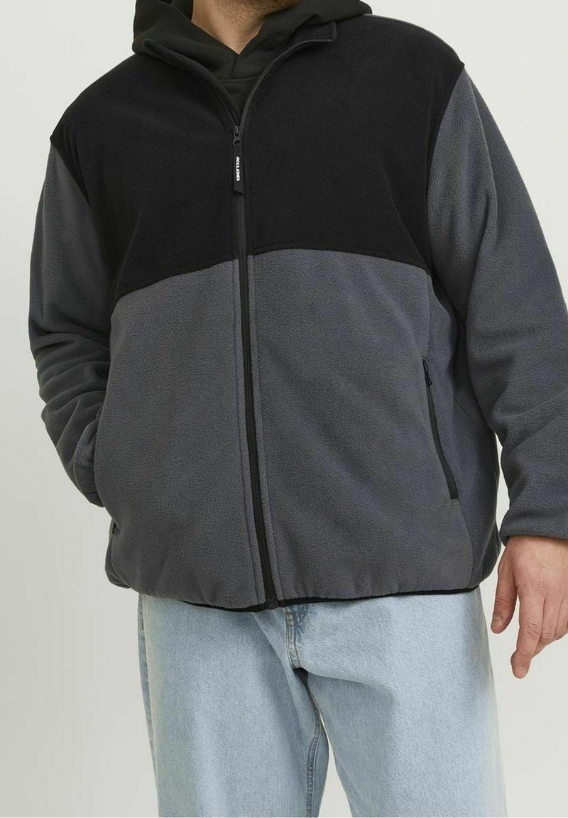 Giacca in pile con sezione superiore nera e sezione inferiore grigia, chiusura a zip, due tasche laterali e vestibilità comoda.