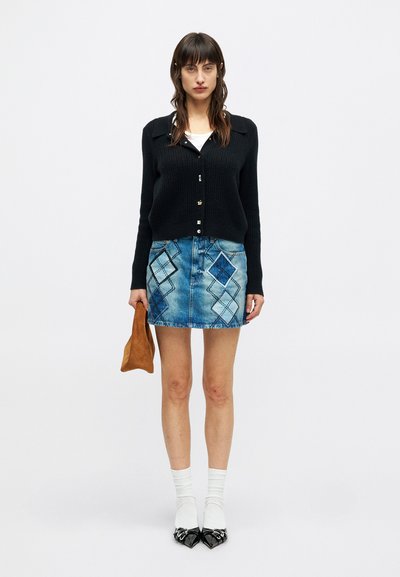 Woman wearing black buttoned cardigan, denim skirt with diamond patches, white socks, black shoes, holding a brown bag, standing against white background.