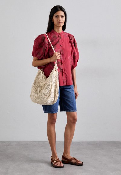 Red floral blouse with puff sleeves, denim shorts, and brown strappy sandals. Holding a large, cream-colored, intricately woven bag.