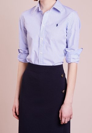 Osoba ubrana w niebiesko-białą koszulę w paski z podwiniętymi rękawami oraz ciemną spódnicę z wysokim stanem z ozdobnymi guzikami.