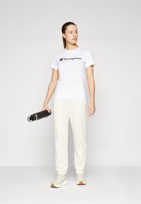 T-shirt blanche avec le logo "Champion", jogging beige clair et baskets crème. Bouteille d'eau noire tenue dans la main droite. Tenue simple et décontractée.