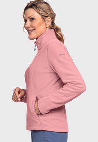 Veste en polaire rose clair avec col montant, fermeture éclair, poches latérales et coutures piquées. Présente un logo sur l'épaule gauche.