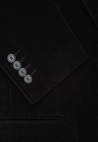 Zbliżenie ciemnej aksamitnej rękawa garnituru z czterema czarnymi guzikami oznaczonymi "Pako Lorente" oraz odpowiadającą kieszeń poniżej.