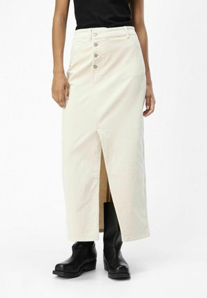 Femme portant une jupe midi crème à boutons sur le devant avec une fente, un haut noir sans manches et des bottines noires, debout sur un fond blanc.