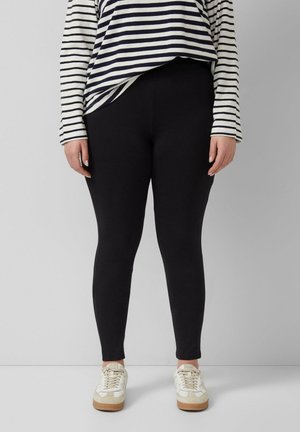 Osoba ubrana w czarne legginsy, białą koszulę z długim rękawem z czarnymi poziomymi paskami oraz białe trampki z jasnobrązowymi podeszwami.