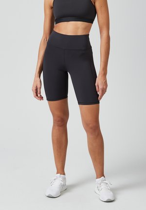 Zwarte high-waisted fietsbroek gemaakt van rekbaar materiaal, met een gladde textuur en minimale naden, gecombineerd met een zwarte sportbeha en witte sneakers.