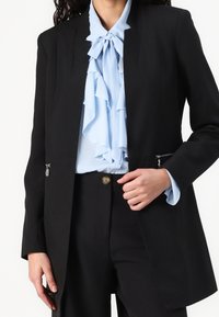 Svart skräddarsydd kavaj med eleganta linjer, försedd med fickor med dragkedja. Under kavajen en ljusblå volangblus med detaljer i form av en slips.