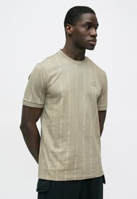 Camiseta beige de manga corta, hecha de algodón, con cuello redondo y un patrón sutil repetido del logo sobre toda la tela.