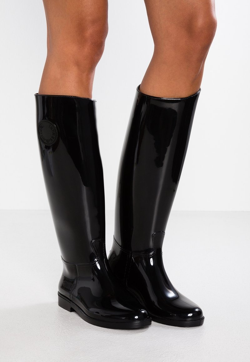 Schwarze gummistiefel in knielanger Ausführung mit glänzender Oberfläche, ausgestattet mit einem runden Logo an der Seite und einer abgerundeten Zehenpartie. Glatte Textur und Knöchel-hohe Sohle.