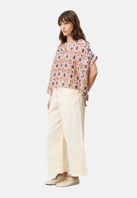 Bunt gemusterte Bluse mit kurzen Ärmeln, kombiniert mit cremefarbenen weiten Hosen und beigen Ballerinas. Das Outfit zeichnet sich durch geometrische und florale Designs aus.