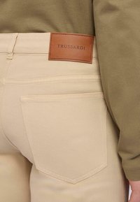 Pantalones ajustados beige con un sutil detalle de costura, que cuentan con una etiqueta de cuero marrón estampada con "TRUSSARDI" en la cinturilla.