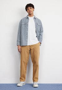 Stribet blå og hvid skjorte med knapper over en hvid T-shirt, parret med khaki bukser og grå sneakers. Simpelt design, afslappet pasform.