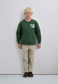 Sudadera verde con bolsillo frontal y gráficos impresos, combinada con pantalones beige y zapatillas marrón oscuro, sobre un fondo neutro.
