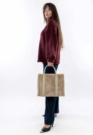 Sac à main en fausse fourrure beige avec des poignées gris clair, en forme rectangulaire, et un détail de logo subtil. Le modèle porte un sweat maroon et un jean foncé.