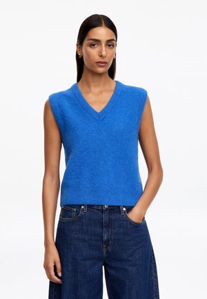 Femme aux cheveux raides et foncés porte un pull sans manches bleu vif à col en V et un jean bleu foncé, debout avec une main dans la poche.