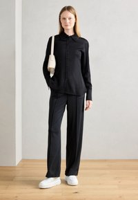 Chemise noire à bouton, pantalon noir ample, et baskets blanches à plateforme. Un sac banane crème complète la tenue. Tissu lisse et coupe ajustée.
