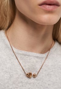 Närbild av en kvinna som bär ett halsband i roséguld med en skivstångsformad hängsmycke över en ljusgrå tröja.