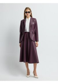 Blazer in vinile bordeaux con taglio sartoriale, abbinato a una gonna a metà lunghezza dello stesso colore. Camicia bianca sottostante, completata da tacchi a punta.