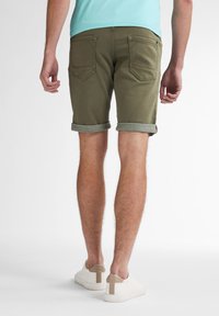 Shorts vert olive avec ourlet retroussé, en tissu coton. Le dos présente deux poches plaquées. Portés avec des baskets blanches avec des accents beiges.